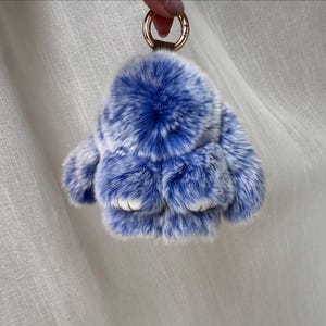 May include: A fluffy blue and white rabbit keychain with a gold-colored ring. The keychain is made of soft, faux fur material. The rabbit has white paws and a round, fluffy body. The keychain is held up by a person's hand.