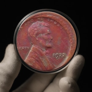 Puede incluir: Un centavo de Lincoln de 1922, en un estuche protector. La moneda muestra un perfil de Abraham Lincoln y el año 1922. La moneda tiene un tono marrón rojizo, con las palabras "IN GOD WE TRUST" y "LIBERTY" visibles.