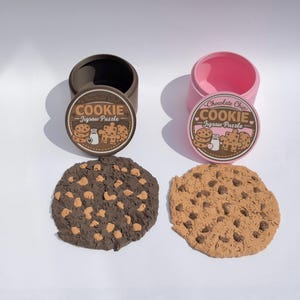 May include: Two round containers, one brown and one pink, each with a lid labelled Chocolate Chip COOKIE Jigsaw Puzzle. Below each lid is a corresponding cookie-shaped jigsaw puzzle, one dark brown with light chips and one tan with dark chips.