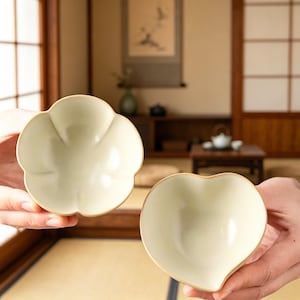 May include: Two ceramic bowls, one flower-shaped and the other heart-shaped, are held up. The bowls have a pale green interior and a light brown rim. The background shows a traditional Japanese interior with a tatami mat.
