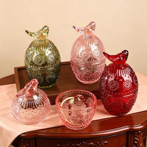 May include: Three egg-shaped glass jars with bird-shaped lids in olive green, light pink, and deep red, featuring embossed floral patterns. A separate light pink glass lid and a matching open bowl containing a beaded bracelet are also present.
