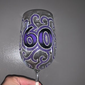 May include: Purple and black wine glass with the number 60 painted on it. The number is surrounded by a white and purple swirl design.