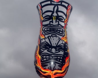 May include: A clear glass tiki mug with a black and white tiki face design and red and yellow flames painted on the side. The mug has a wide base and a narrow top.