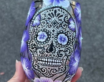 May include: A clear glass with a hand-painted sugar skull design in black, white, and silver. The skull is surrounded by purple flowers.