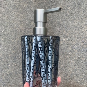 May include: A clear glass soap dispenser with a silver pump. The dispenser has a black and white design of birch trees.