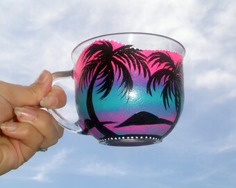 May include: A clear glass mug with a hand-painted design of palm trees and a sunset. The sunset is painted in shades of pink, purple, blue, and green.