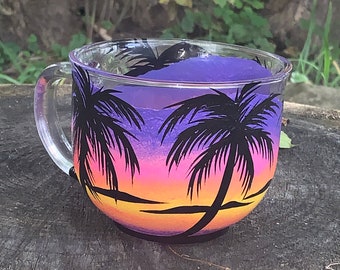 May include: A glass mug with a painted design of palm trees and a sunset in shades of purple, pink, orange, and yellow.