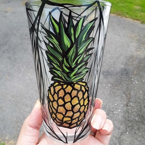 May include: A clear glass with a black and white geometric design featuring a hand-painted pineapple with green leaves and a yellow body.