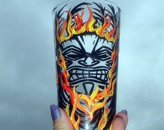 May include: A clear glass cup with a black and white tiki face design and yellow and red flames painted on the side.