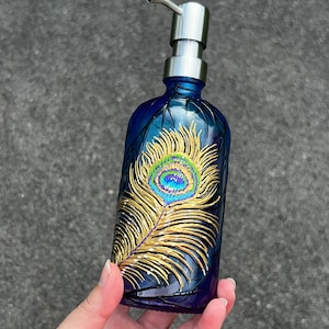 May include: Blue glass soap dispenser with a peacock feather design painted on the front. The feather is gold with a blue, green, and purple eye.