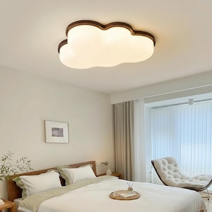 May include: A cloud-shaped ceiling light fixture with a brown trim and a white, glowing center. The light is mounted on a white ceiling in a bedroom setting. The room features a bed with white bedding, a wooden headboard, and a comfortable armchair.