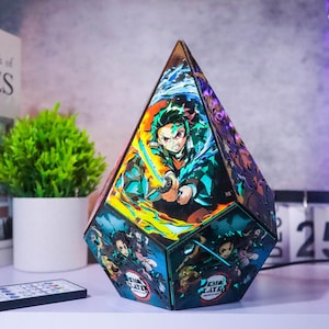 May include: A multi-faceted, illuminated decorative lamp featuring anime-style artwork. The lamp has a geometric shape with images of characters from "Demon Slayer" on its sides. The lamp is lit with various colors and includes a remote control.