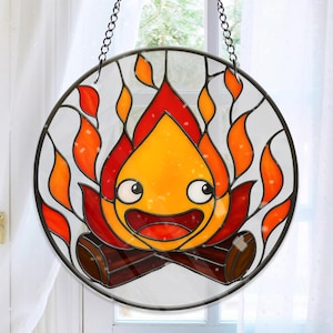 May include: A round stained glass window featuring a cartoon campfire character with a smiling face. The flames are in shades of red, orange, and yellow, with brown logs at the base. The window is framed in black and hangs from a chain.