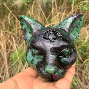 May include: A carved stone cat head figurine featuring a third eye on the forehead. The sculpture is crafted from a polished mineral with a mottled pattern of deep forest green and dark magenta-red hues, showcasing a smooth, glossy surface finish.