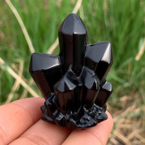 May include: A cluster of polished black obsidian crystals. The crystals have sharp, geometric shapes and a glossy surface. The base of the cluster is textured, and the crystals are held in a hand against a blurred green background.