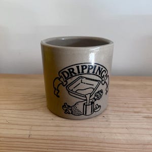 May include: A light beige ceramic crock featuring a black graphic print. The design includes the word DRIPPING in a banner above an illustration of a square pan pouring liquid into a small container, surrounded by two small mushroom icons.