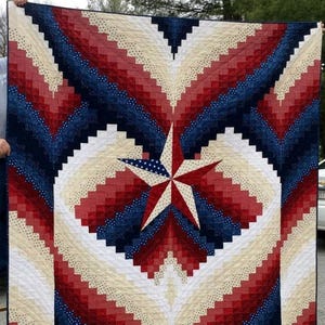 May include: A patriotic quilt featuring a central star design. The quilt showcases a pattern of red, white, and blue fabrics arranged in a starburst pattern. The star is made of red, white, and blue fabrics. The quilt is held up by a wooden stand.