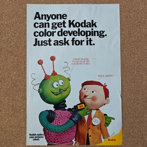 May include: A vintage Kodak advertisement poster featuring a green robot with red antennae and a person in a tan suit. The text reads: Anyone can get Kodak color developing. Just ask for it. I WANT KODAK TO DEVELOP MY COLOR PICTURES. SPACE SHOTS? Kodak makes your pictures count.