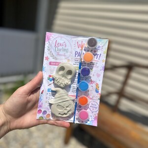May include: A craft kit featuring two unpainted plaster figures, a skull and a pumpkin wearing a hat, packaged with a small paintbrush and a strip of six circular paint pots in brown, orange, purple, blue, orange, and pink on a printed card.