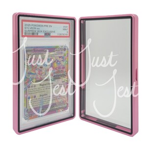 May include: A pink protective case, open to reveal a Pok&eacute;mon trading card. The card features a Sylveon ex design with the text "Magical Charm" and "Prismatic Evolutions". The case has a black border and the text "2025 POKEMON PRE EN SYLVEON ex SURPRISE BOX EXCLUSIVE".