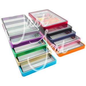 May include: A collection of colorful card cases with clear tops. The cases are in various colors, including red, black, blue, pink, orange, green, purple, and silver. One case contains a trading card. The cases are stacked and arranged.
