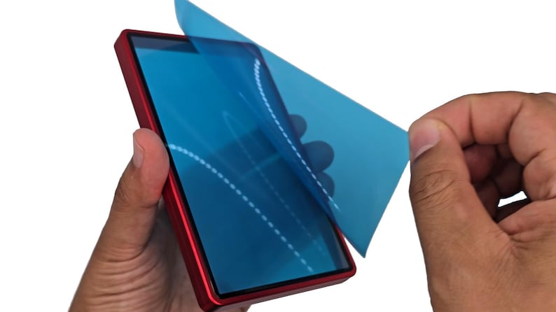 May include: A red smartphone with a blue screen protector being peeled off. The phone is held by two hands against a white background. The screen protector has a dotted design.