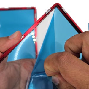 May include: A red frame with a blue protective film being peeled away. The frame has a white interior and is held by two hands. Additional frames with blue film are visible in the background.