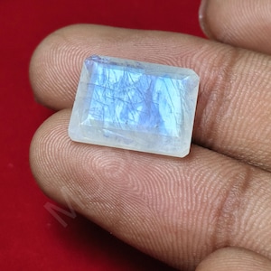 May include: A rectangular, faceted moonstone gemstone with a translucent, milky white appearance and a distinct blue adularescence or sheen across its surface. The stone features visible internal inclusions and a polished, flat top surface.