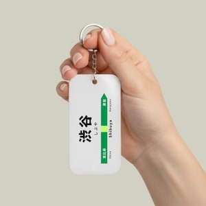 May include: White rectangular keychain with a silver keyring. The keychain features Japanese text and a green line indicating Shibuya station, with Ebisu and Harajuku as adjacent stations. The keychain is held by a person.