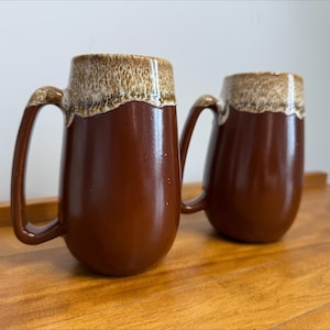 Peut inclure: Deux mugs en céramique marron avec un bord et une anse crème. Les mugs ont une finition brillante et sont posés sur une surface en bois. Les mugs sont conçus avec une grande anse pour une prise en main facile.