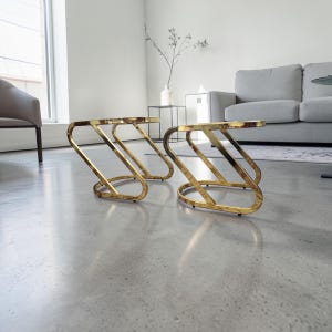 May include: Two gold side tables with a distinctive, curved design. The tables have a circular top and a zig-zag base. They are positioned on a grey concrete floor in a modern living room setting. The tables are approximately 45 cm high.