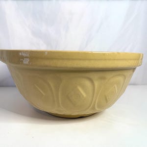 May include: A large, light yellow mixing bowl with a wide rim and embossed floral and diamond patterns. The bowl is made of ceramic and has a slightly worn appearance, suggesting it is vintage or antique.