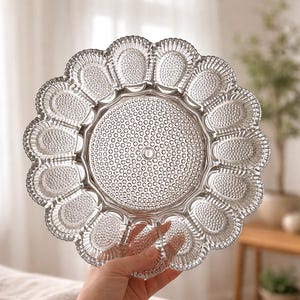 May include: Clear glass serving platter with a scalloped edge and a central circular design. The platter has individual egg-shaped indentations around the edge and a textured surface. A decorative piece for serving appetizers or desserts.