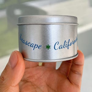 May include: A cylindrical silver metal tin with a lid. The side of the tin features blue cursive text that reads Seascape and California, separated by a small green asterisk symbol. The container has a smooth, metallic finish and a simple design.