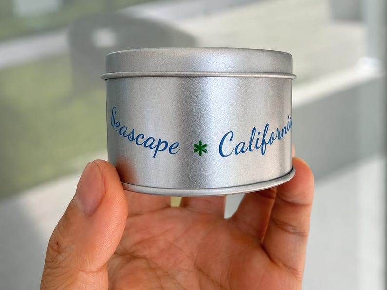 May include: A cylindrical silver metal tin with a lid. The side of the tin features blue cursive text that reads Seascape and California, separated by a small green asterisk symbol. The container has a smooth, metallic finish and a simple design.