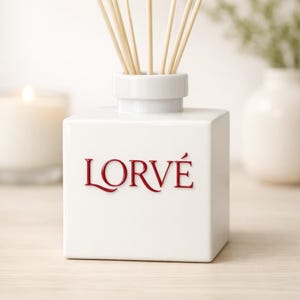 May include: A white square ceramic reed diffuser bottle with a cylindrical neck. The word LORV&Eacute; is embossed in red lettering on the front. Several light-colored wooden reeds extend upward from the opening of the bottle.