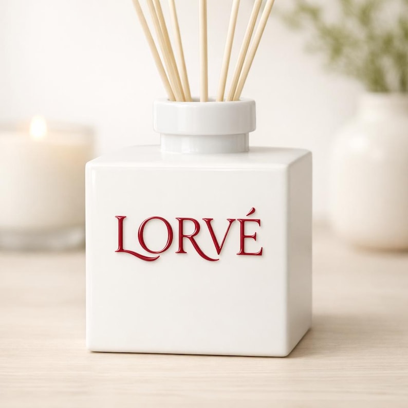 May include: A white square ceramic reed diffuser bottle with a cylindrical neck. The word LORV&Eacute; is embossed in red lettering on the front. Several light-colored wooden reeds extend upward from the opening of the bottle.