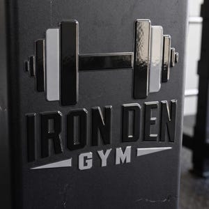 May include: A black metal gym equipment frame featuring a raised, glossy logo. The logo displays a stylized barbell icon above the text IRON DEN in bold black letters, with the word GYM positioned below in silver, flanked by two silver triangles.