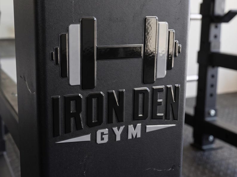 May include: A black metal gym equipment frame featuring a raised, glossy logo. The logo displays a stylized barbell icon above the text IRON DEN in bold black letters, with the word GYM positioned below in silver, flanked by two silver triangles.