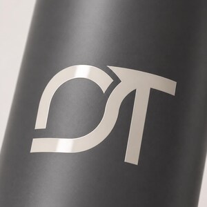 May include: A matte black cylindrical object featuring a large, silver-toned metallic logo consisting of the letters C and T intertwined. The letters are stylized with clean, sharp lines and a reflective finish that contrasts against the dark surface.