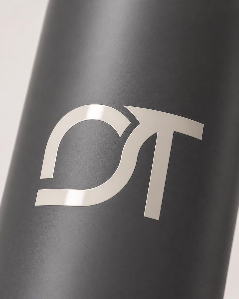 May include: A matte black cylindrical object featuring a large, silver-toned metallic logo consisting of the letters C and T intertwined. The letters are stylized with clean, sharp lines and a reflective finish that contrasts against the dark surface.