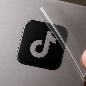 May include: A square black sticker featuring the silver TikTok logo icon with rounded corners. The sticker is shown being applied to a gray surface, with a clear adhesive transfer film partially peeled back from the top right corner of the decal.