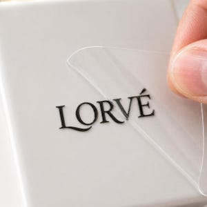 May include: A clear adhesive sticker featuring the black text LORVÉ in a serif font. The sticker is partially peeled back from a smooth, light gray surface, revealing the transparent material and the black lettering printed on the adhesive layer.
