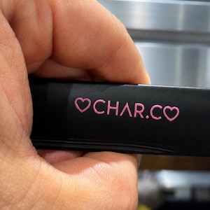 May include: A matte black rectangular object featuring the pink text CHAR.CO flanked by two pink heart symbols. The surface is smooth and dark, providing a high contrast for the bright pink lettering and heart icons printed on the front side.