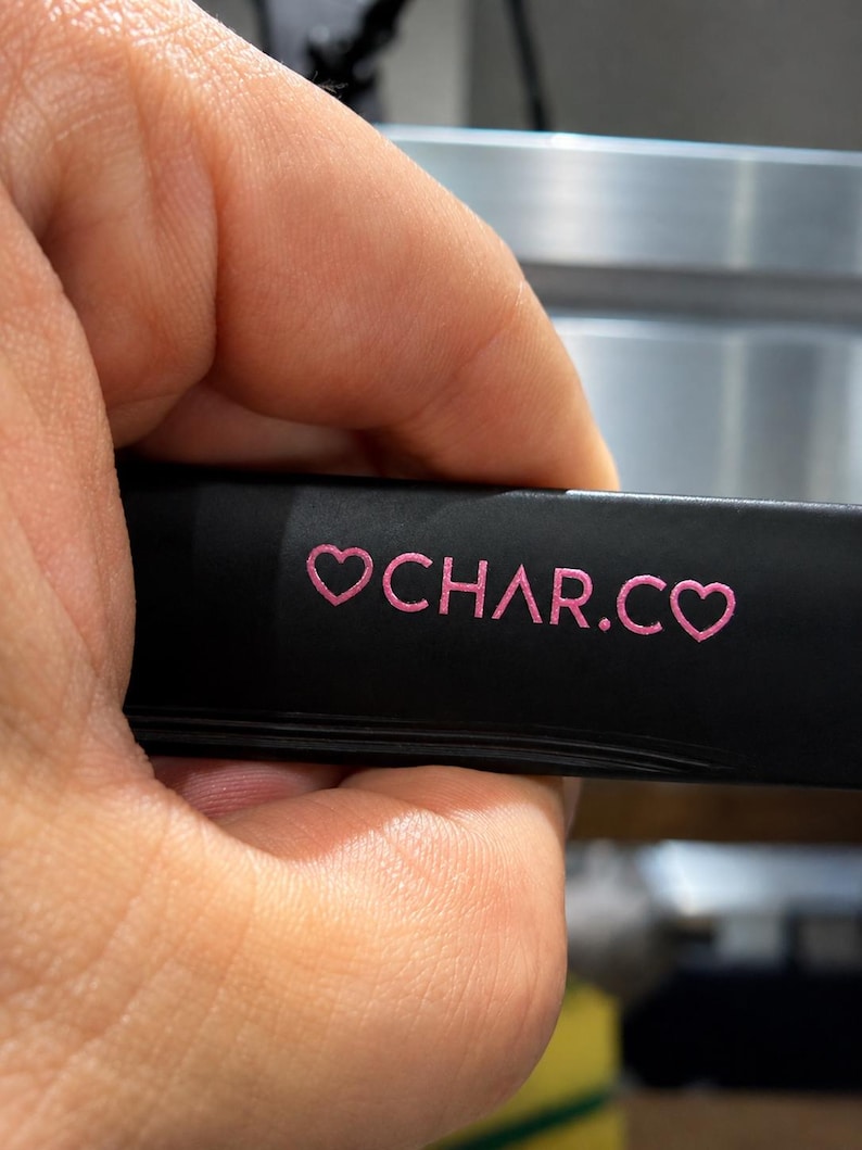 May include: A matte black rectangular object featuring the pink text CHAR.CO flanked by two pink heart symbols. The surface is smooth and dark, providing a high contrast for the bright pink lettering and heart icons printed on the front side.