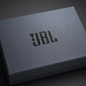 May include: A rectangular dark grey box with a matte finish featuring the JBL logo embossed in a glossy black finish on the top centre. The box has sharp, clean edges and a solid construction, presenting a sleek and uniform appearance throughout.