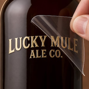 May include: A dark brown glass bottle featuring a clear adhesive label with the gold-colored text LUCKY MULE ALE CO. printed on it. The label is partially peeled back from the surface of the bottle, revealing the glossy finish of the dark glass.