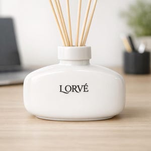 May include: A white ceramic reed diffuser bottle with a wide, oval-shaped base and a narrow neck. The word LORV&Eacute; is printed in black on the front. Several light-colored wooden reeds are inserted into the top opening of the diffuser vessel.