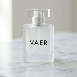 May include: A rectangular perfume bottle made of frosted glass with the word VAER printed in black on the front. The bottle features a clear, rectangular cap and a silver-toned spray nozzle mechanism visible beneath the transparent lid.