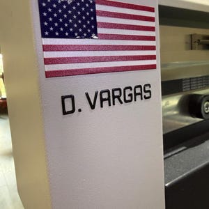 May include: A white machine surface featuring a rectangular sticker of the United States flag positioned above the black text D. VARGAS. The machine has a smooth, matte finish and a curved edge, with metallic components visible in the background.
